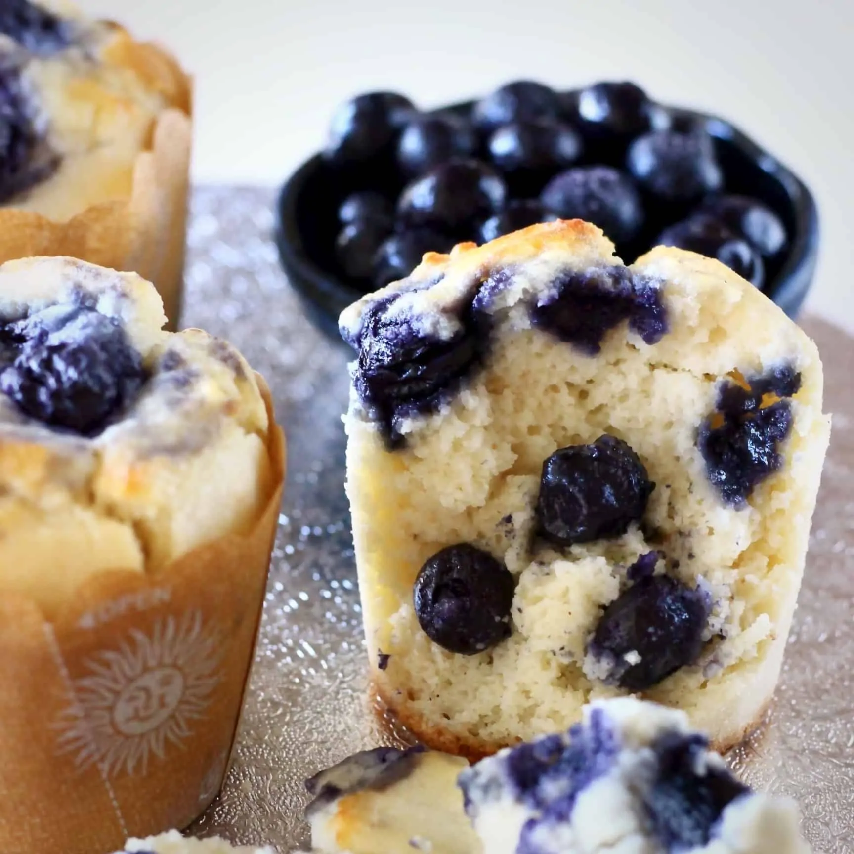 Three vegan blueberry muffins with a bite taken out of one with a bowl of fresh blueberries in the background