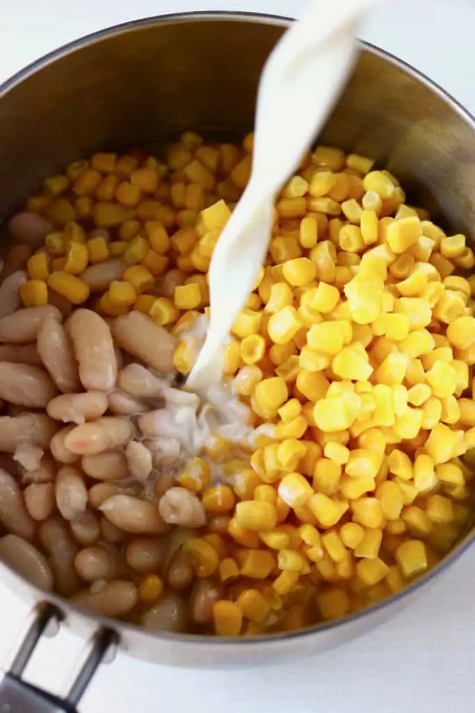 White beans and sweetcorn in a silver saucepan with milk being poured in