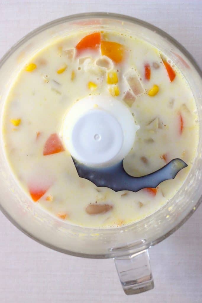 Sweetcorn, milk, white beans and vegetables in a food processor against a white background