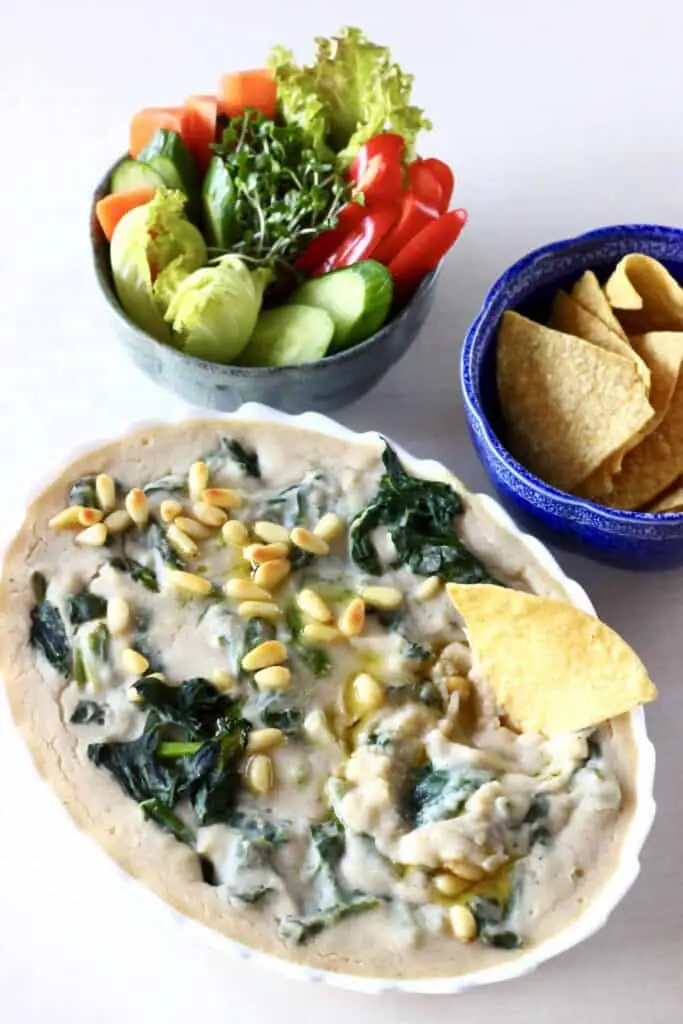 Photo of an oval baking dish filled with creamy spinach and artichoke dip topped with pine nuts with a bowl of tortilla chips and a bowl of vegetable crudités with a tortilla chip sticking into the dip