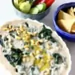 Photo of an oval baking dish filled with creamy spinach and artichoke dip topped with pine nuts with a bowl of tortilla chips and a bowl of vegetable crudités