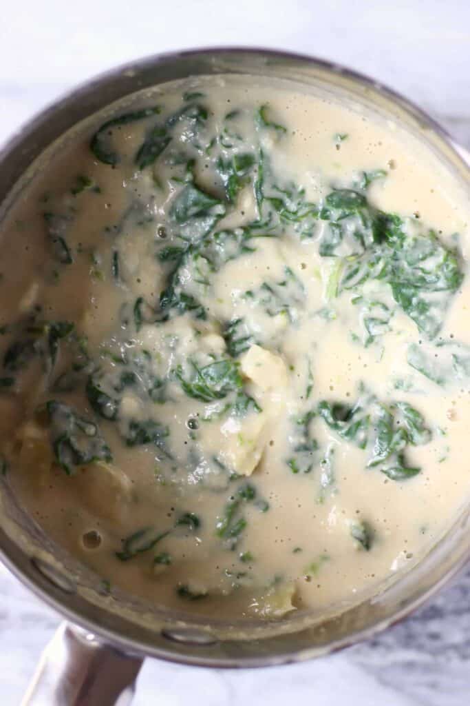 Photo of a white sauce in a silver saucepan with chopped artichokes and cooked spinach