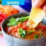 A blue bowl of orange vegan quest topped with green pepper and chopped coriander on a pink background with chopped vegetables and a dark blue bowl of tortilla chips with a tortilla chip being dipped into the quest