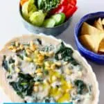 Photo of an oval baking dish filled with creamy spinach and artichoke dip topped with pine nuts with a bowl of tortilla chips and a bowl of vegetable crudités
