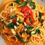 Spaghetti in a creamy red sauce topped with green spinach, sliced red peppers and pine nuts on a dark brown plate against a marble background