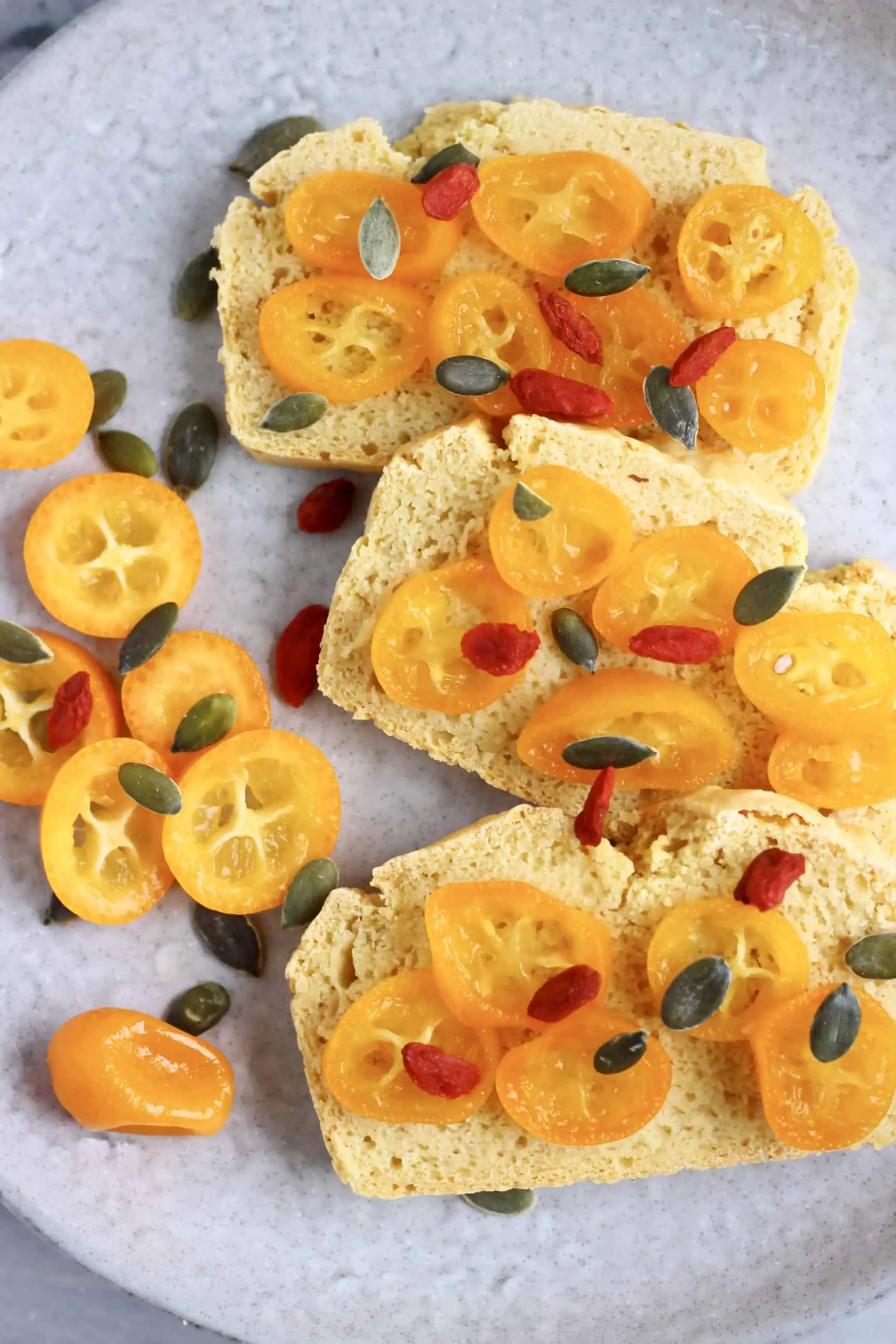 Three slices of gluten-free vegan bread topped with sliced kumquats, goji berries and pumpkin seeds on a grey plate