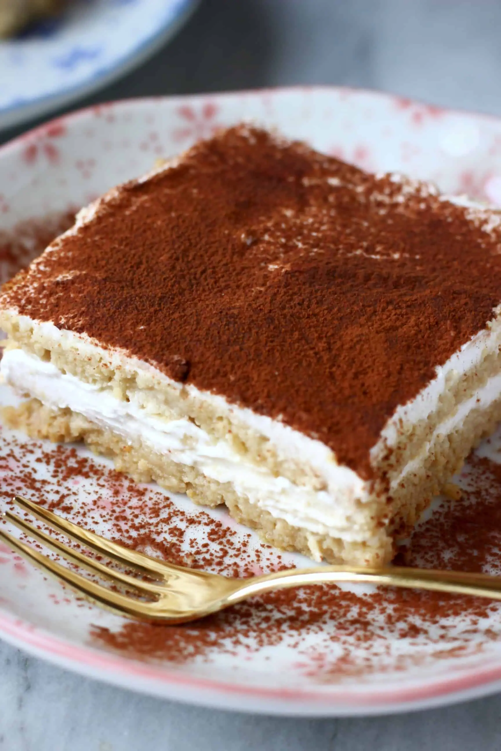 A square of gluten-free vegan tiramisu with a bite taken out of it on a white plate with pink flowers and a gold fork