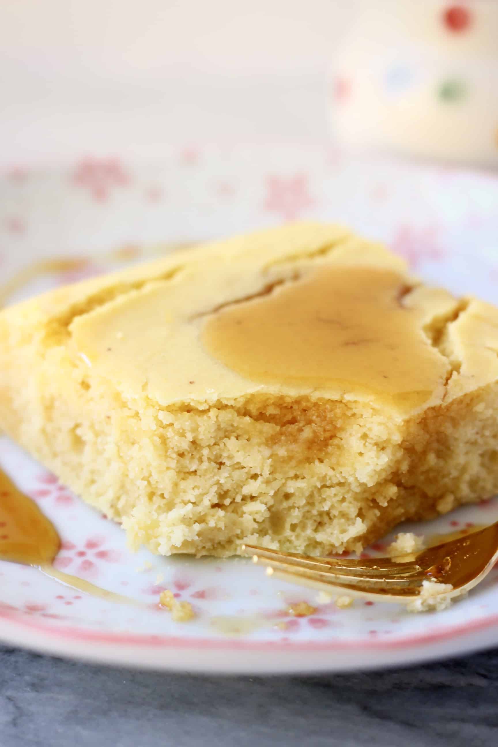 A square of gluten-free vegan cornbread with a bite taken out of it drizzled with maple syrup on a plate with a fork