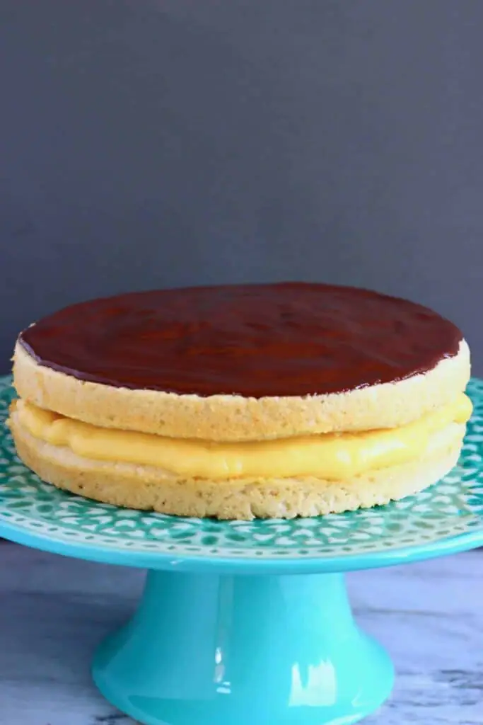 A golden brown sponge cake filled with yellow custard topped with chocolate ganache on a green cake stand against a grey background