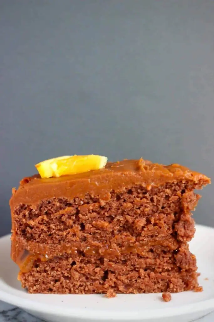 Photo of a slice of chocolate cake topped with an orange wedge on a white plate against a grey background