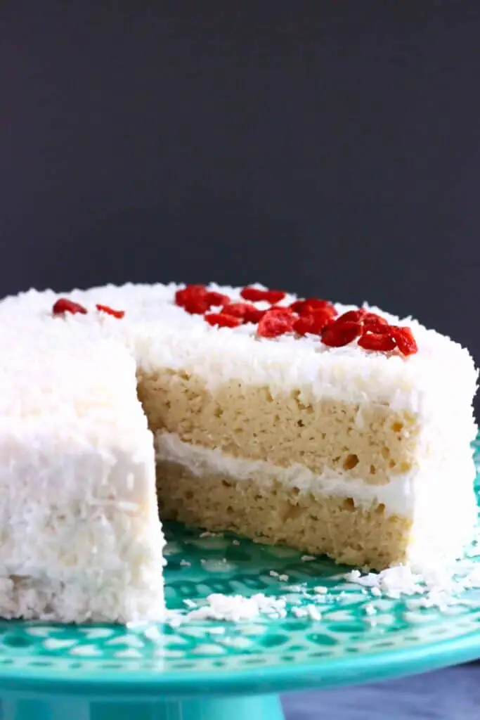 Photo of a sliced sponge cake frosted with creamy white frosting, coated in coconut and topped with red goji berries on a green cake stand against a dark grey background