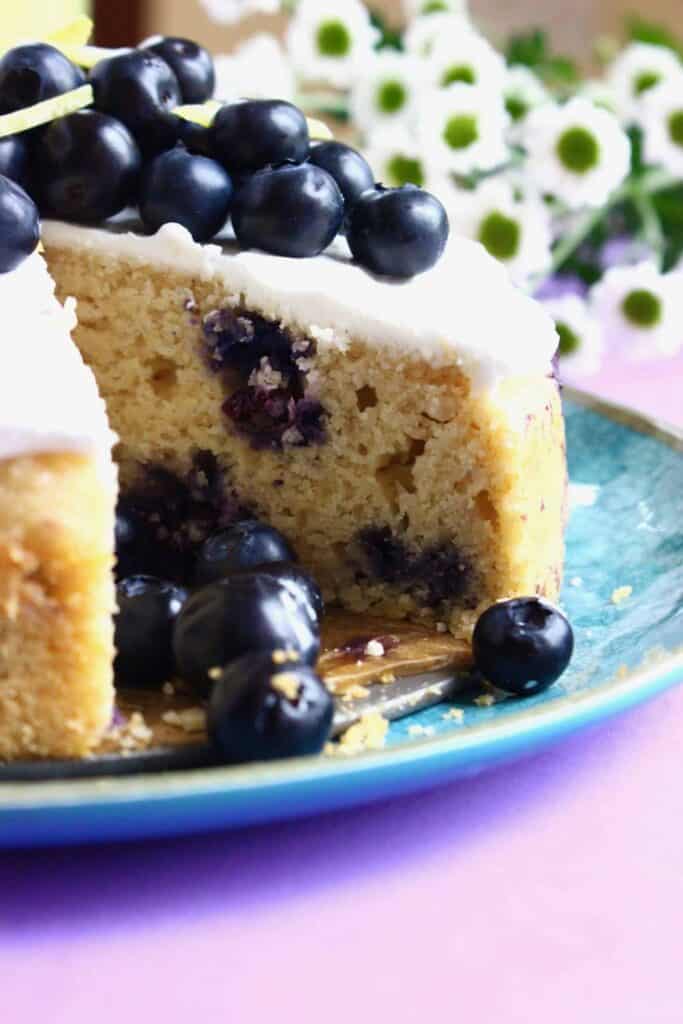 Photo of a sliced sponge cake with blueberries in it topped with white creamy frosting and fresh blueberries on a blue plate against a purple background with small white flowers