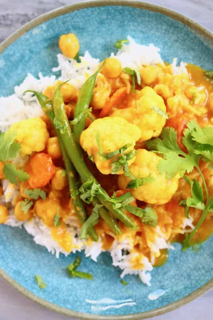 Cauliflower and green beans in an orange curry sauce on a pile of white rice on a blue plate against a marble background