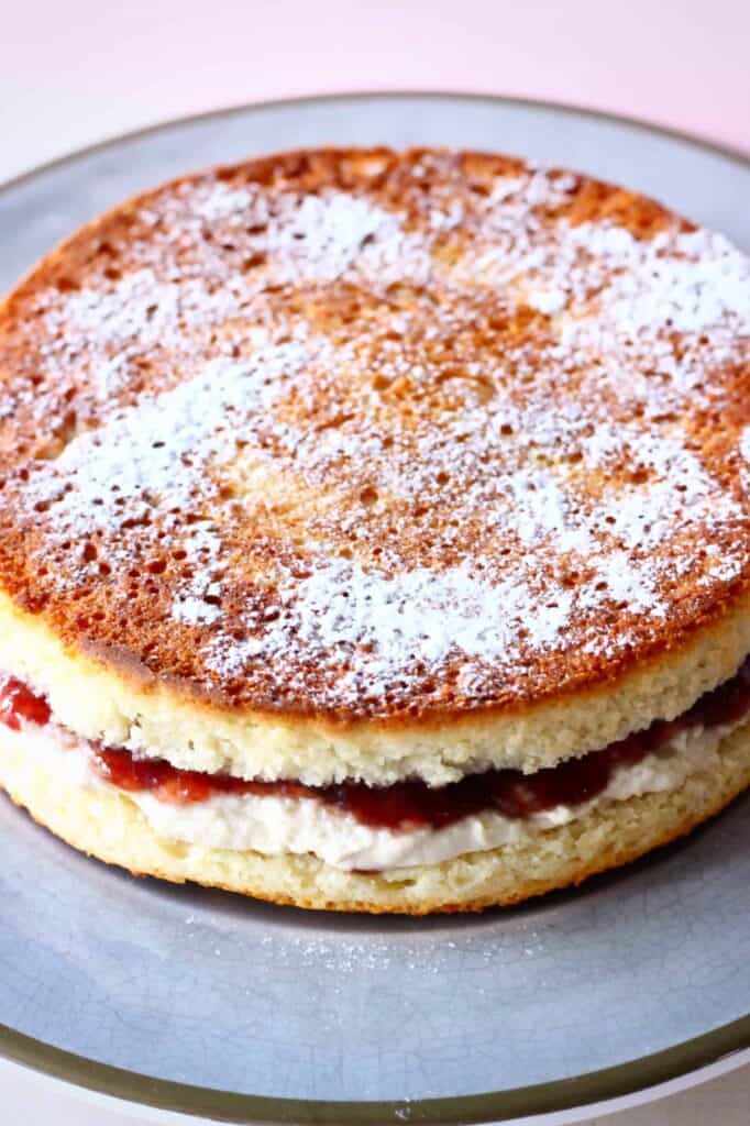 Photo of a golden brown sponge cake sandwiched with strawberry jam and white buttercream dusted with icing sugar on a grey plate
