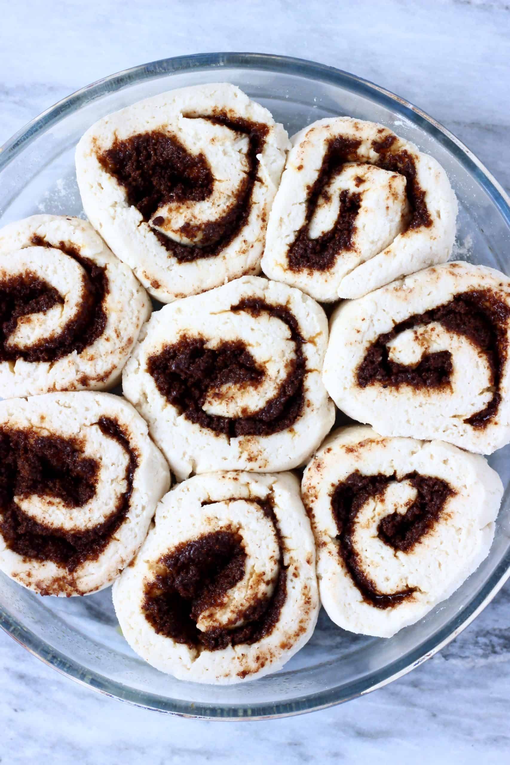 Eight raw gluten-free vegan cinnamon rolls in a round baking dish that have doubled in size after proving