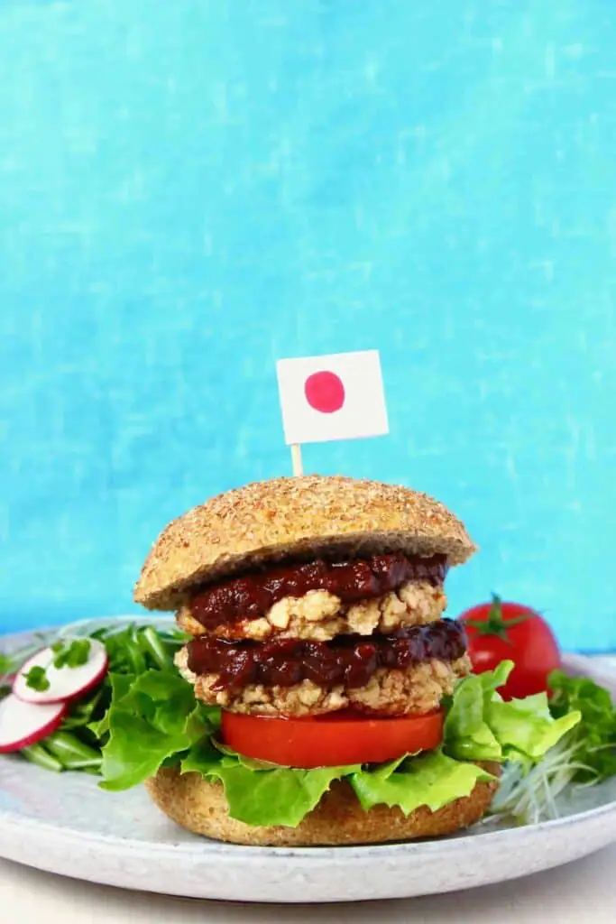 A tofu burger with brown sauce, tomato and lettuce on a grey plate with a Japanese flag on a toothpick sticking out of it against a bright blue background