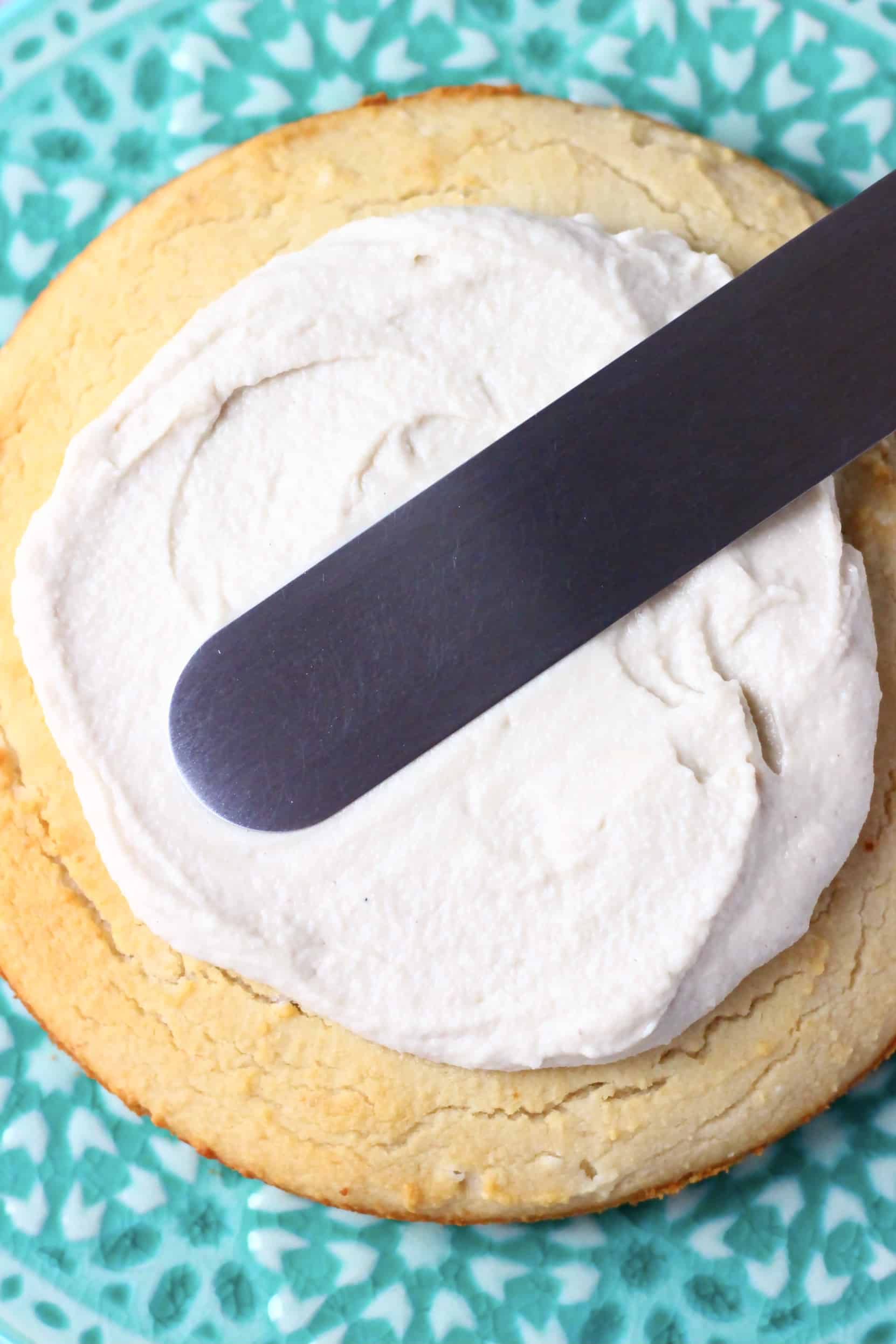 A gluten-free vegan vanilla cake sponge on a cake stand being spread with vegan buttercream frosting using a palette knife
