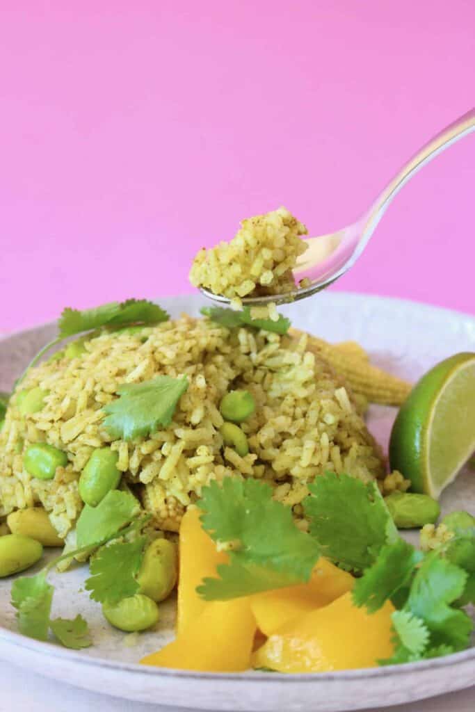 A pile of green rice on a plate with a silver spoon holding up a mouthful of it against a pink background