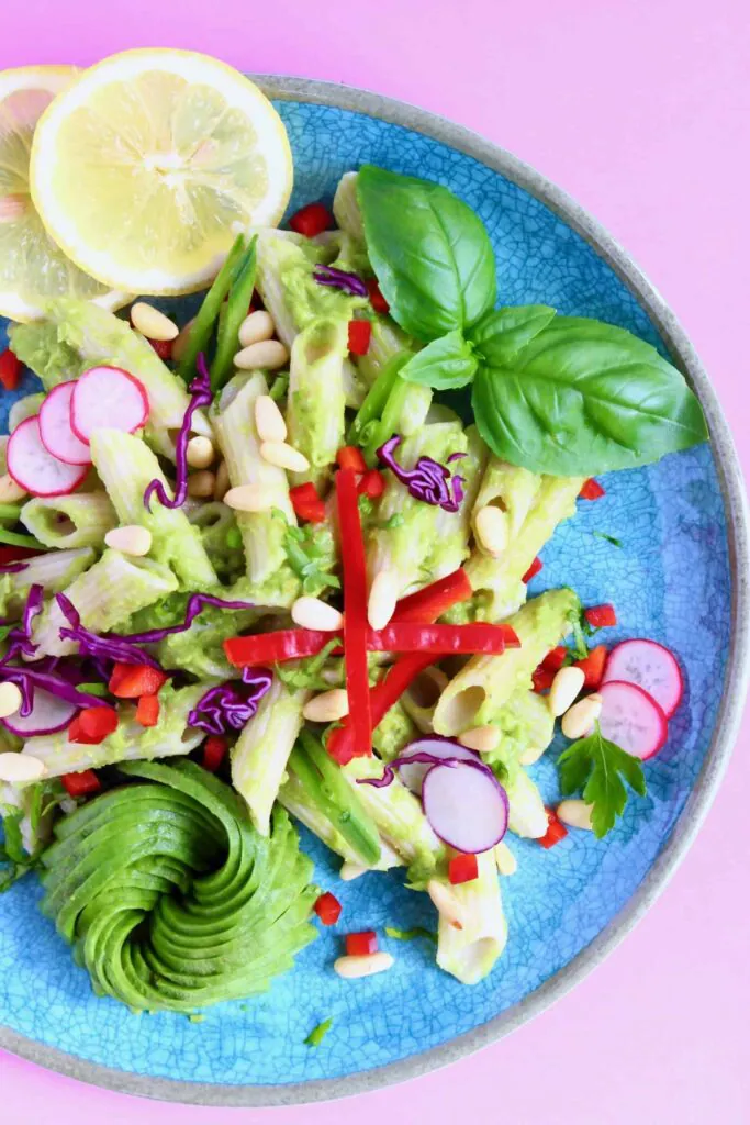 Penne pasta in a green avocado sauce with vegetables on a blue plate against a pink background