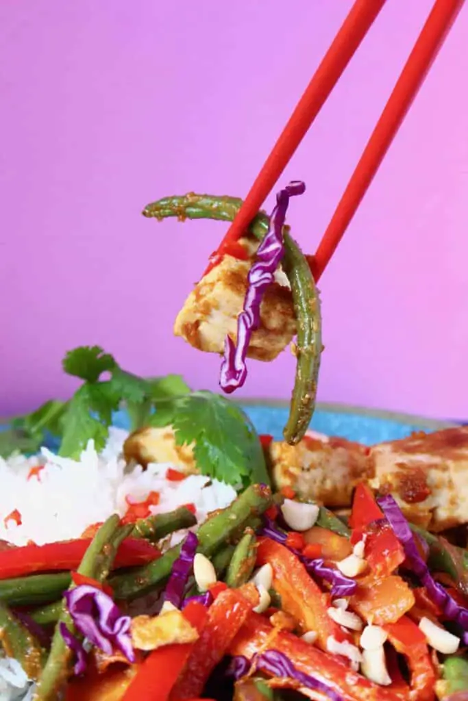 A tofu and vegetable stir fry on a plate with a pair of red chopsticks holding up a mouthful against a pink background