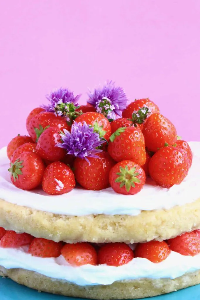 Photo of a sponge cake frosted with white creamy frosting topped with a pile of tiny strawberries and purple flowers against a pink background
