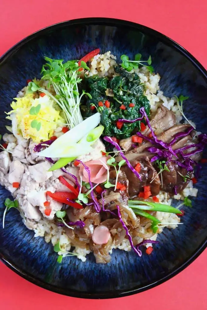 Scrambled tofu, oyster mushrooms, spinach, green cress and other vegetables on a bed of rice in a dark blue bowl against a red background