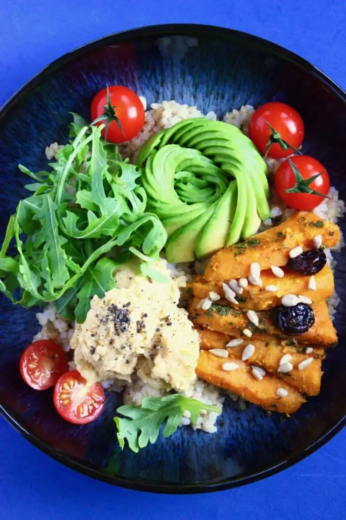 Sweet potato sticks, an avocado rose, rocket, hummus and cherry tomatoes on a bed of rice in a dark blue bowl against a dark blue background