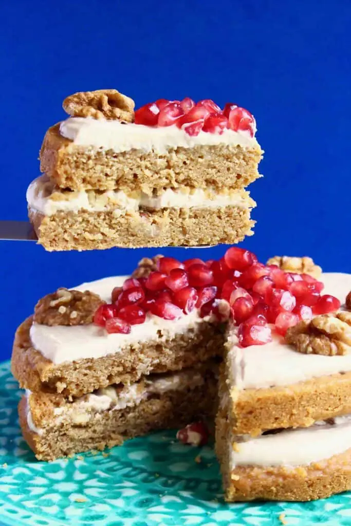 Photo of a coffee cake sandwiched with creamy white frosting and topped with walnuts and pomegranate seeds with one slice being held up in the air against a dark blue background