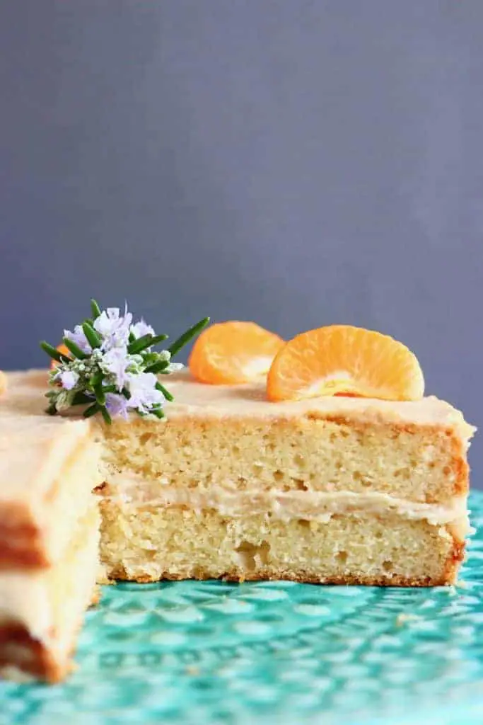 A sliced sponge cake with buttercream frosting topped with orange segments and a sprig of rosemary on a green cake stand against a grey background