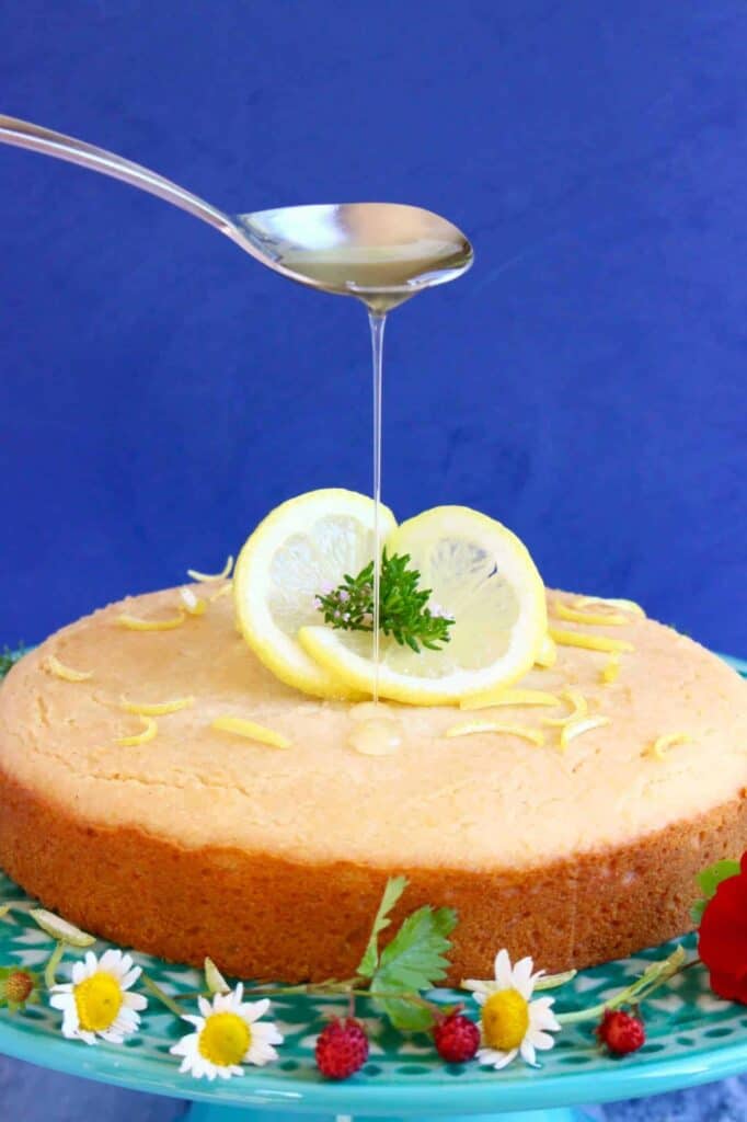 Photo of a cake topped with a lemon slice with a spoon drizzling syrup over it on a green cake stand covered in flowers against a dark blue background