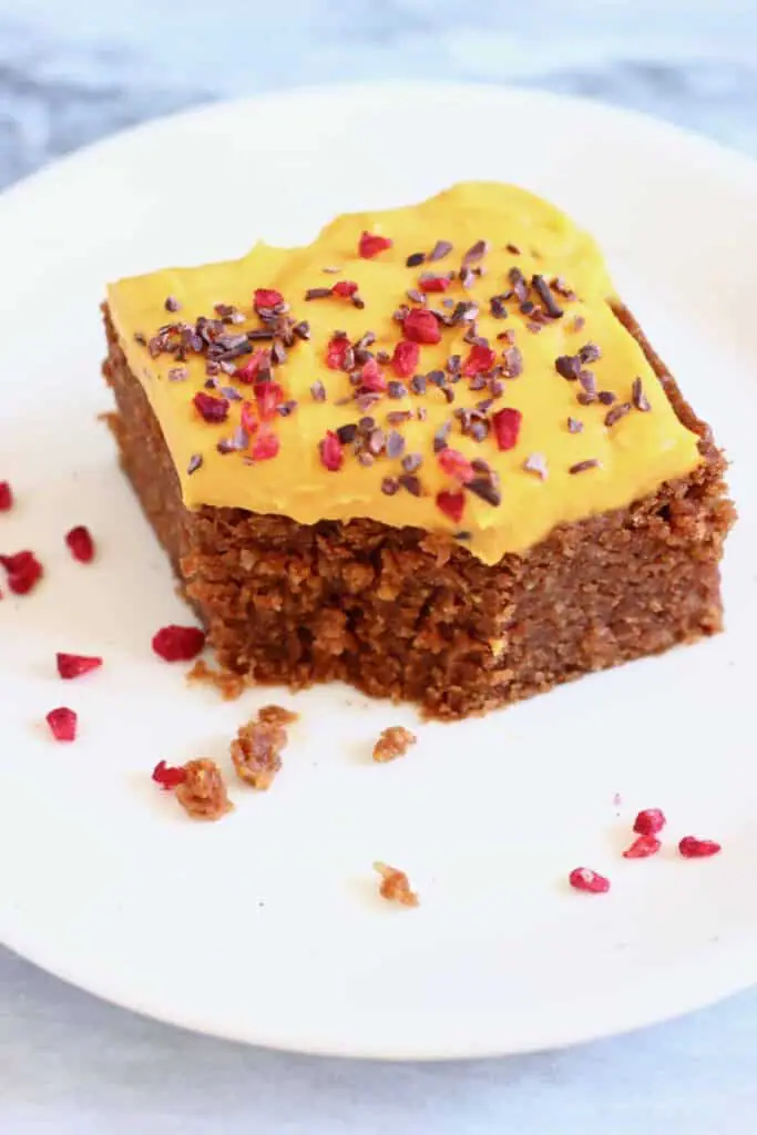 Photo of a square slice of chocolate cake topped with yellow frosting sprinkled with cacao nibs and freeze-dried raspberries on a small white plate against a marble background