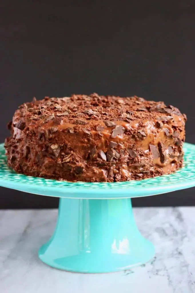 Photo of a chocolate cake covered in bits of chopped up chocolate on a blue cake stand against a grey background