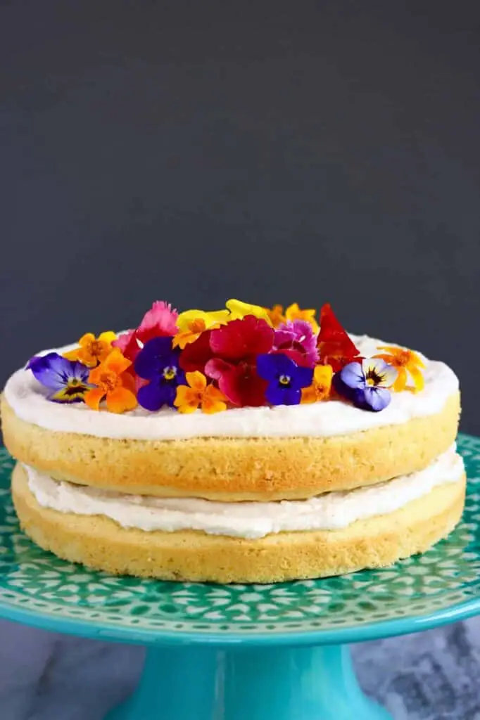 Photo of a yellow sponge cake with white creamy frosting topped with edible flowers on a blue cake stand against a grey background