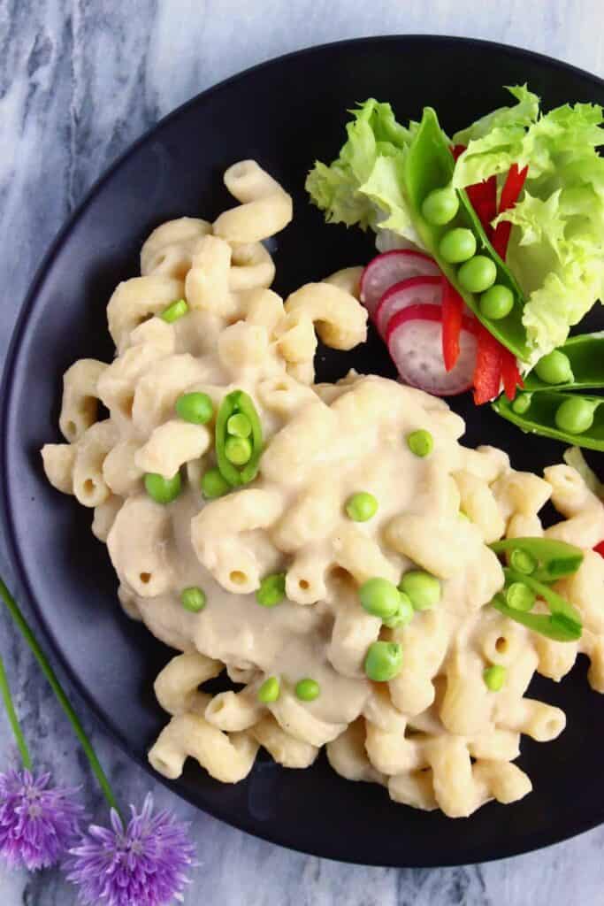 Mac and cheese with green peas on a black plate with salad against a marble background