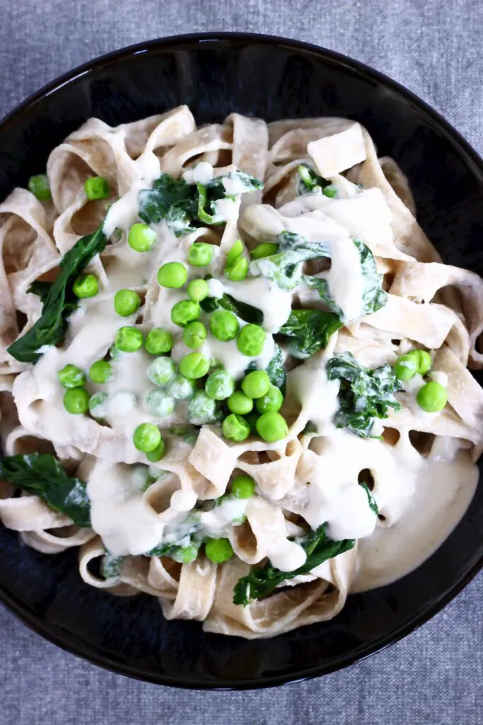Tagliatelle in a white cream sauce with spinach and green peas in a black bowl against a grey background