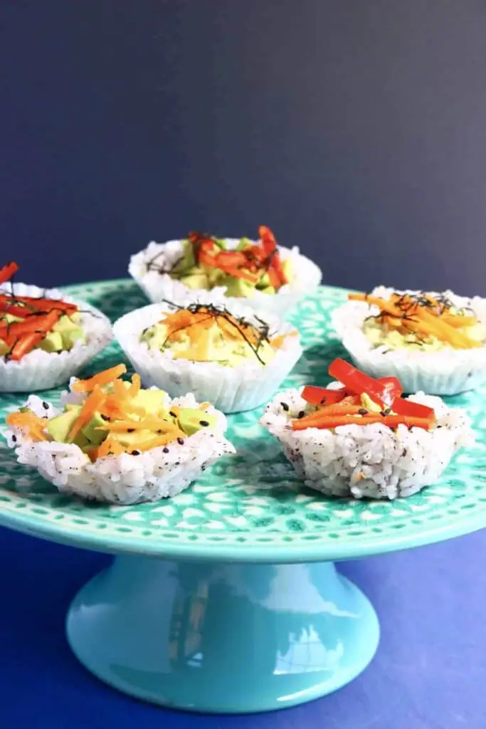 Six muffin-shaped sushi topped with grated carrot and shredded red pepper on a green cake stand against a grey background