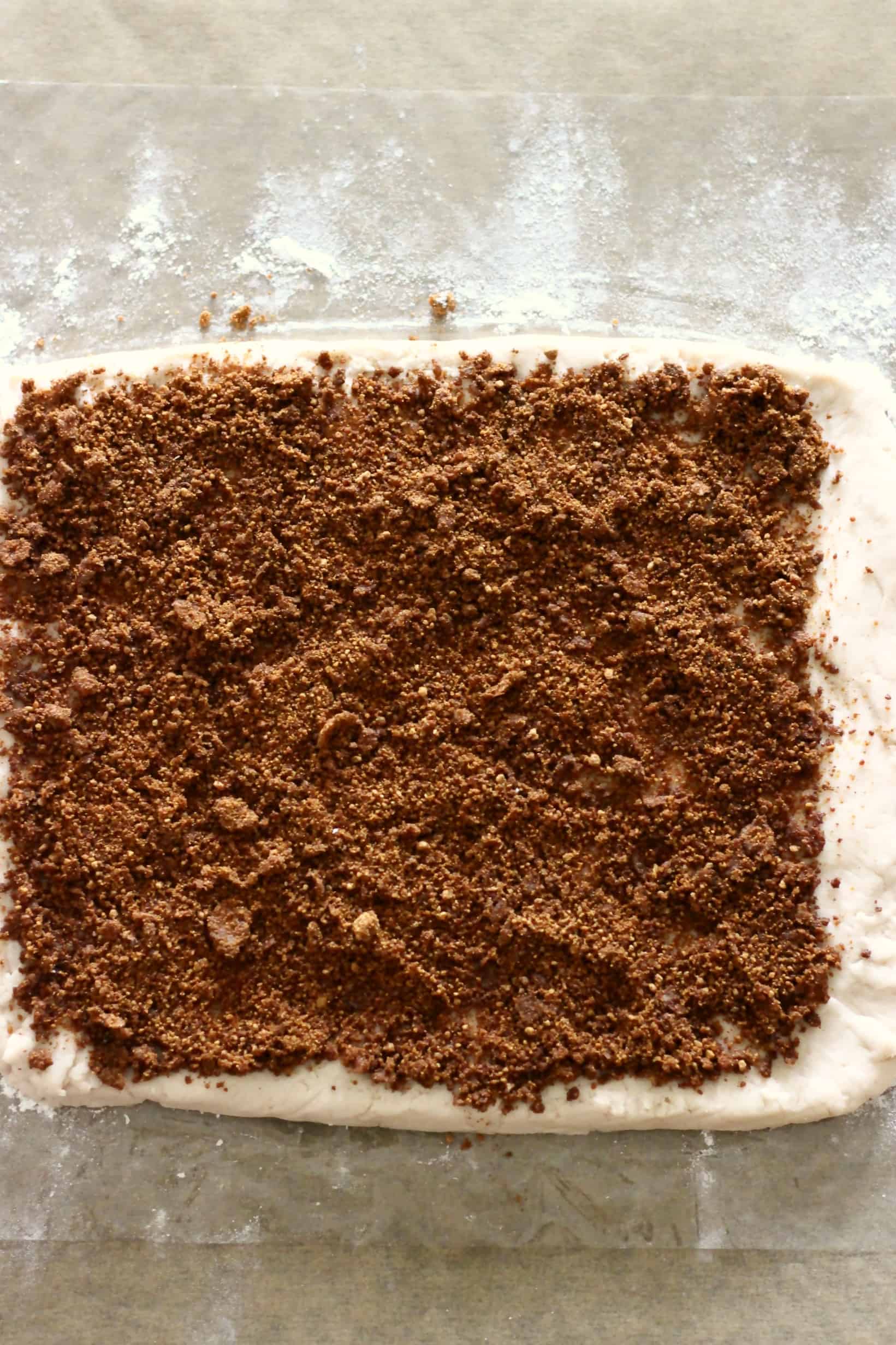 A square of gluten-free vegan cinnamon roll dough sprinkled with cinnamon sugar on a piece of cling film sprinkled with flour on a sheet of baking paper