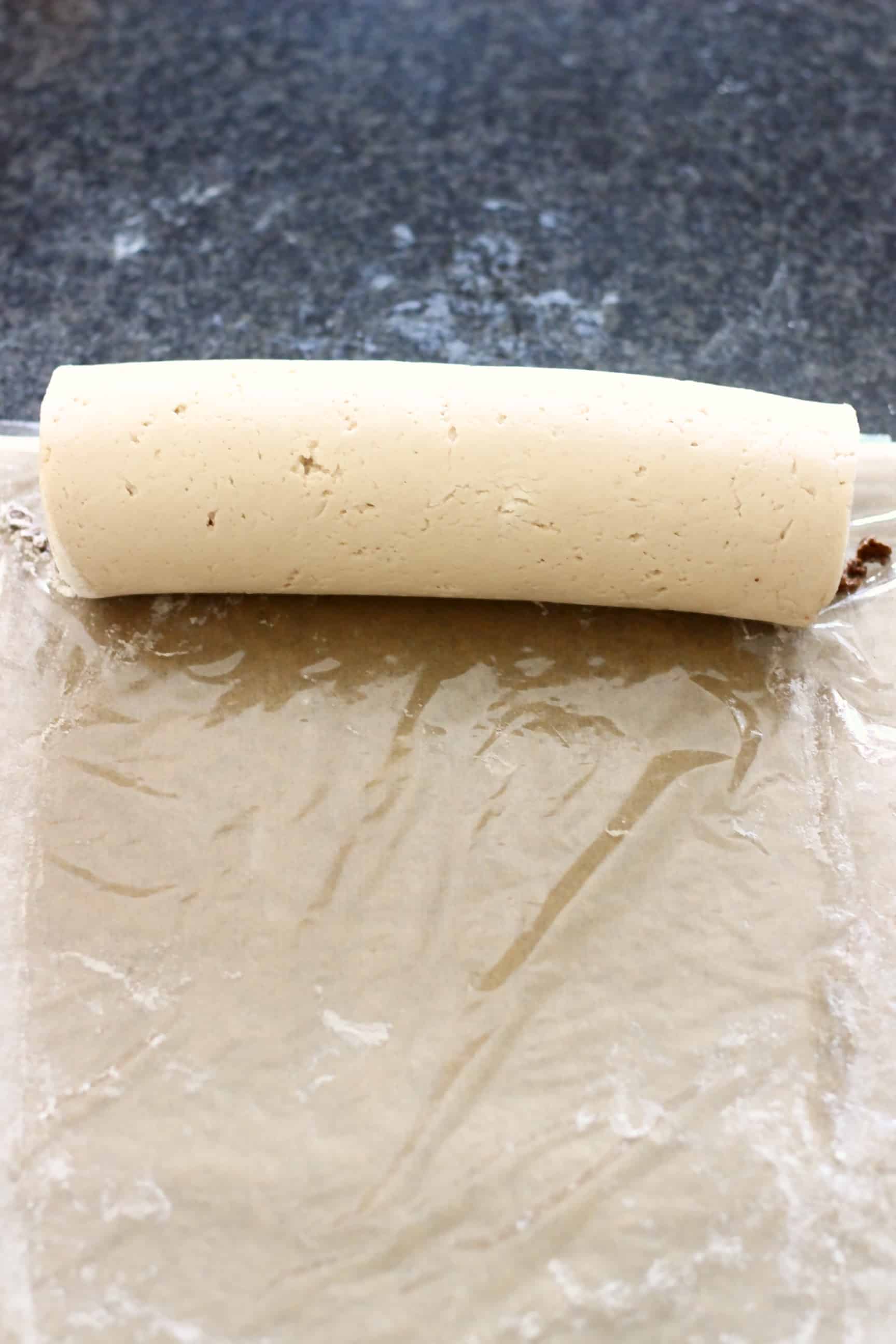 Gluten-free vegan cinnamon roll dough filled with cinnamon sugar rolled into a sausage shape on a piece of cling film on baking paper