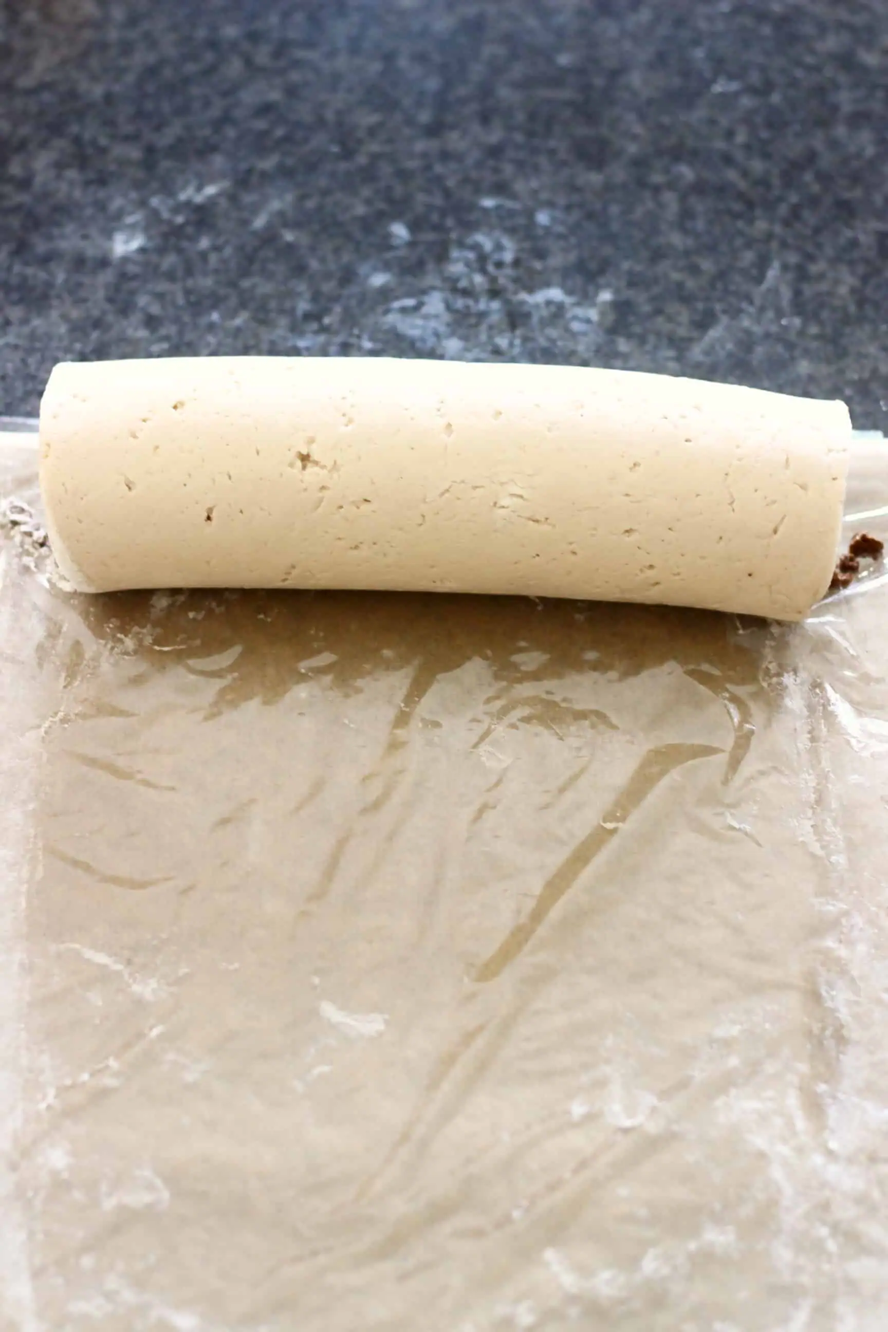 Gluten-free vegan cinnamon roll dough filled with cinnamon sugar rolled into a sausage shape on a piece of cling film on baking paper