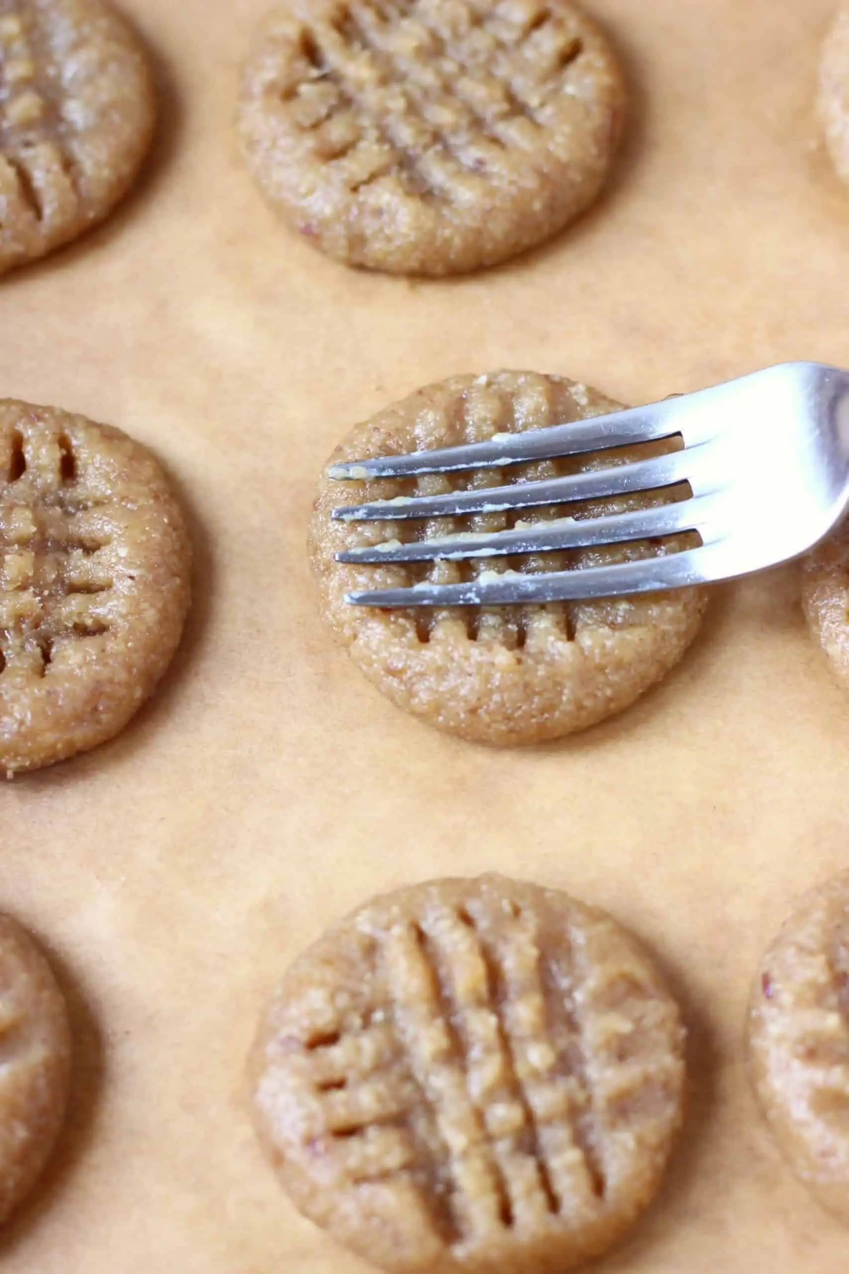 Eight raw gluten-free vegan peanut butter cookies on a baking tray lined with baking paper with a fork making criss-cross patterns on top