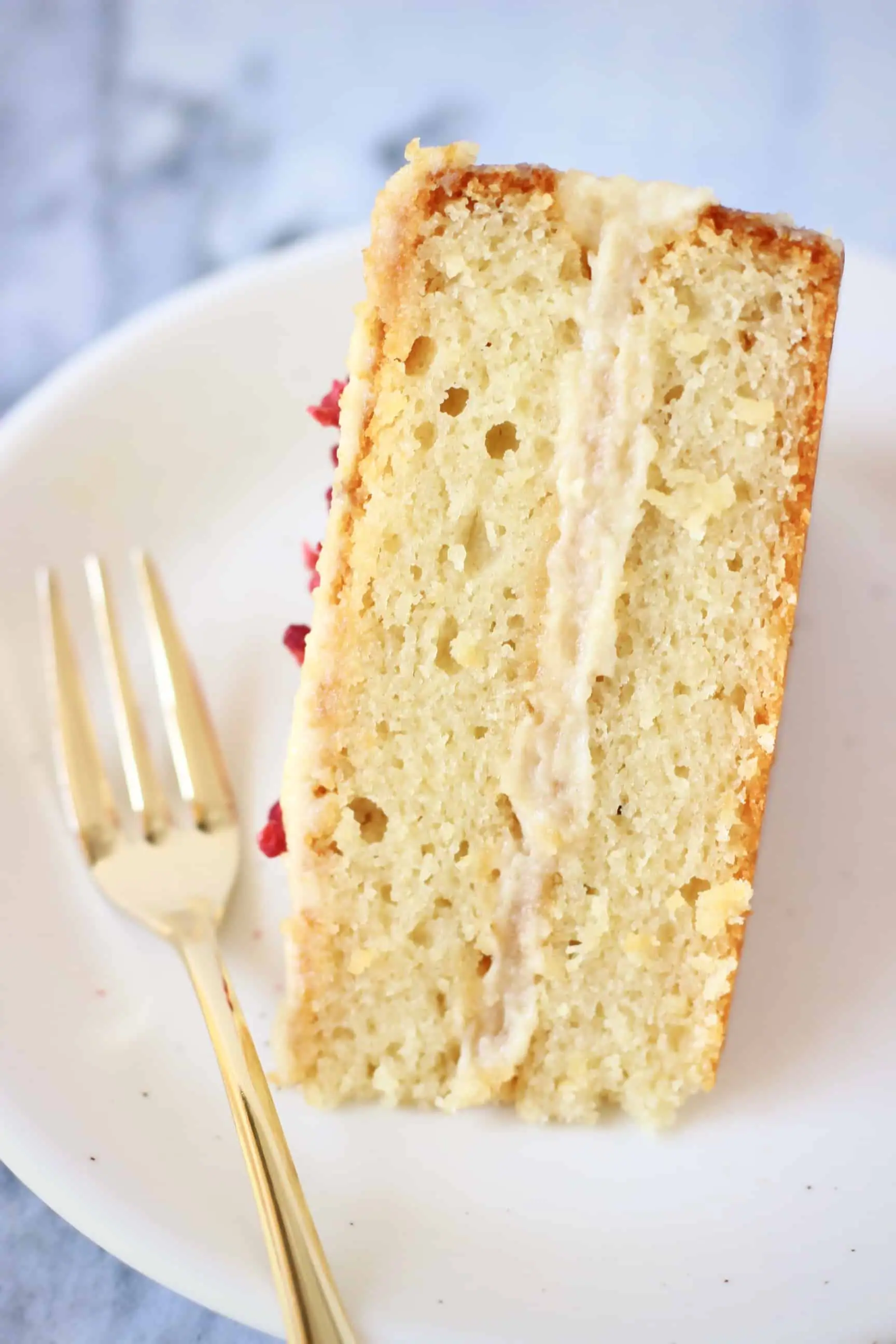 A slice of gluten-free vegan vanilla  cake with white frosting on a plate with a gold fork