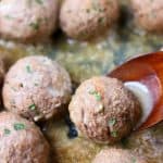 Brown meatballs in a brown gravy topped with green herbs with a wooden spoon lifting up one meatball