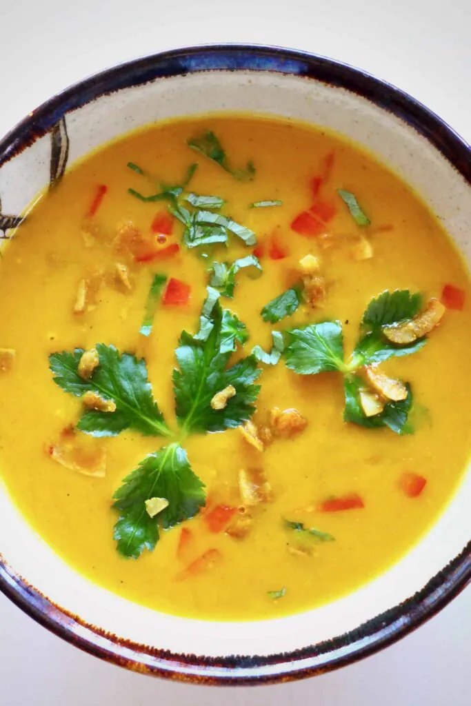 Photo of orange soup topped with coconut flakes, red peppers and green herbs in a white bowl with a dark brown rim