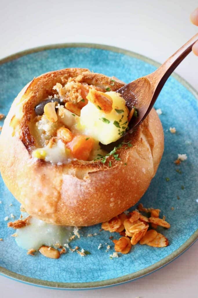 Photo of a white vegetable stoup in a bread bowl on a blue plate with a wooden spoon holding up a mouthful of soup