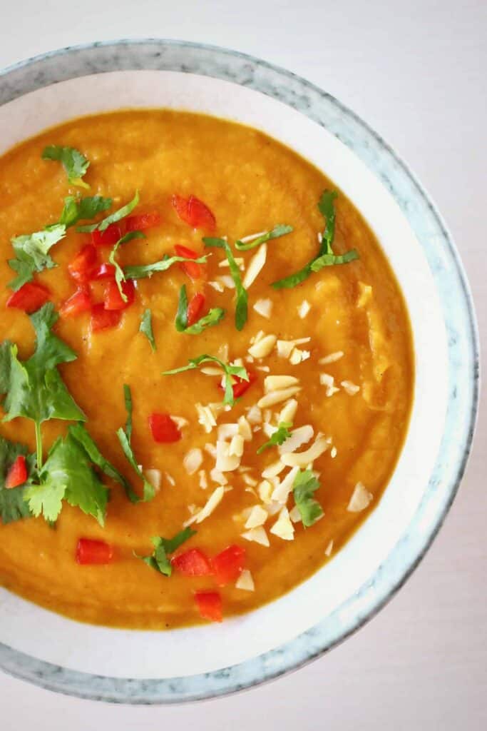Photo of orange soup topped with green herbs, red peppers and chopped peanuts in a grey bowl against a white background 