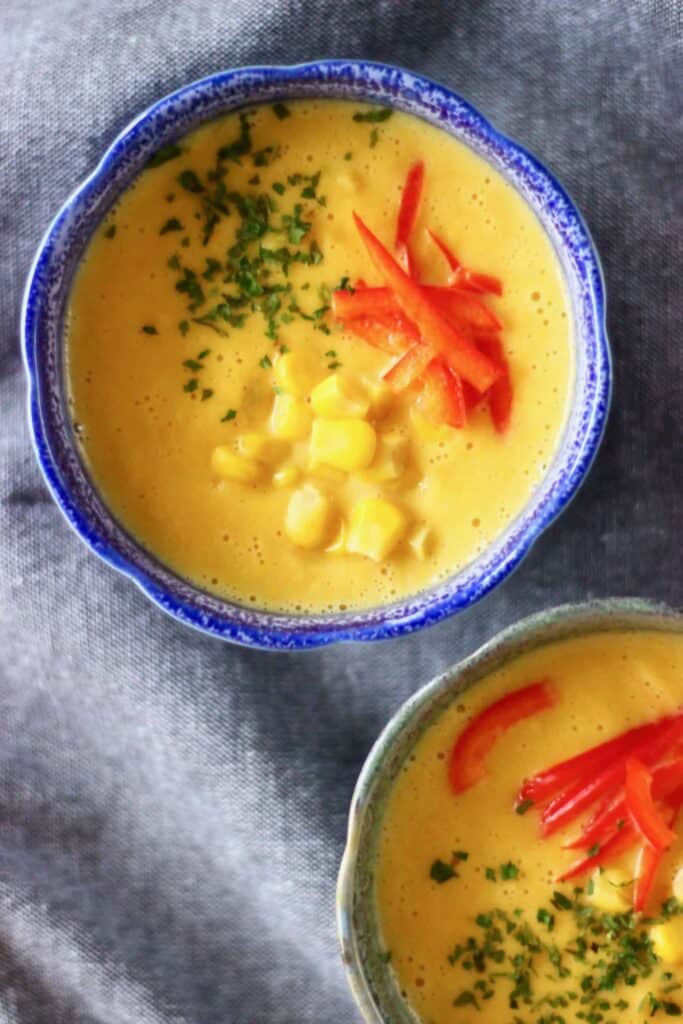 Photo of two bowls of yellow soup topped with yellow sweetcorn and red peppers and green herbs against a grey fabric background