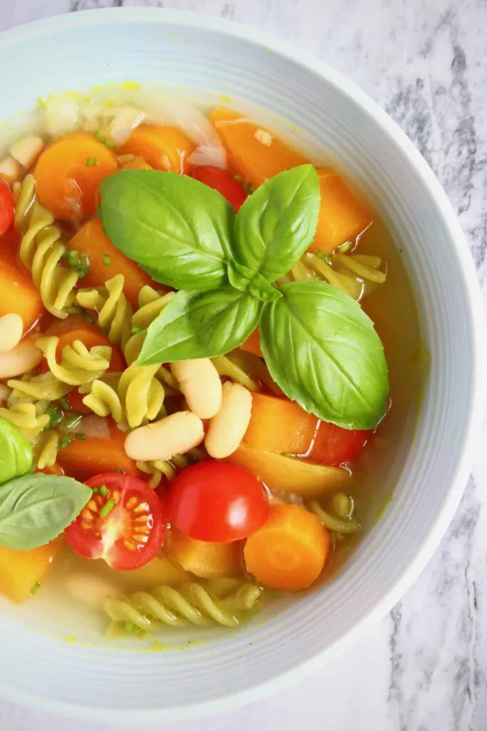 Soup with green pea pasta, carrots, white beans, tomatoes and a sprig of basil in a light blue bowl against a marble background
