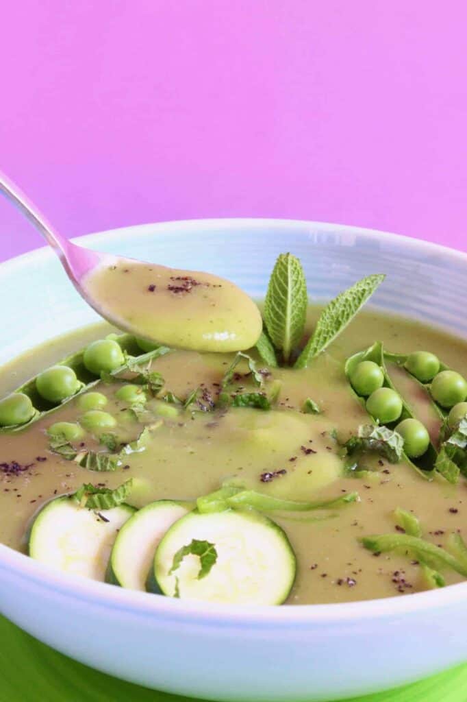 A green soup topped with sliced zucchini, green peas, black pepper and a mouthful of soup being held up with a silver spoon against a pink background