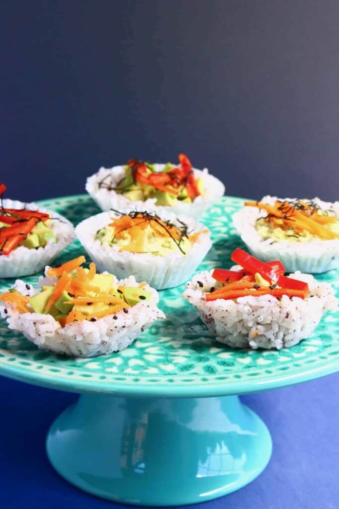 Six muffin-shaped sushi topped with grated carrot and shredded red pepper on a green cake stand against a grey background