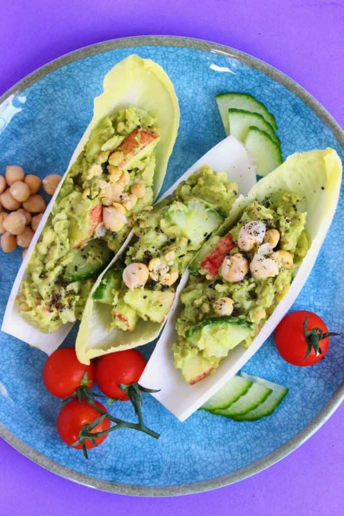 Three chicory boats filled with avocado, chickpeas, apples and cucumber on a blue plate against a purple background